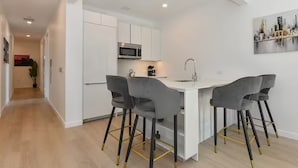 A modern kitchen with white cabinets, a central island with bar stools, and a microwave. - Suites at Dog Lane (Mansfield)