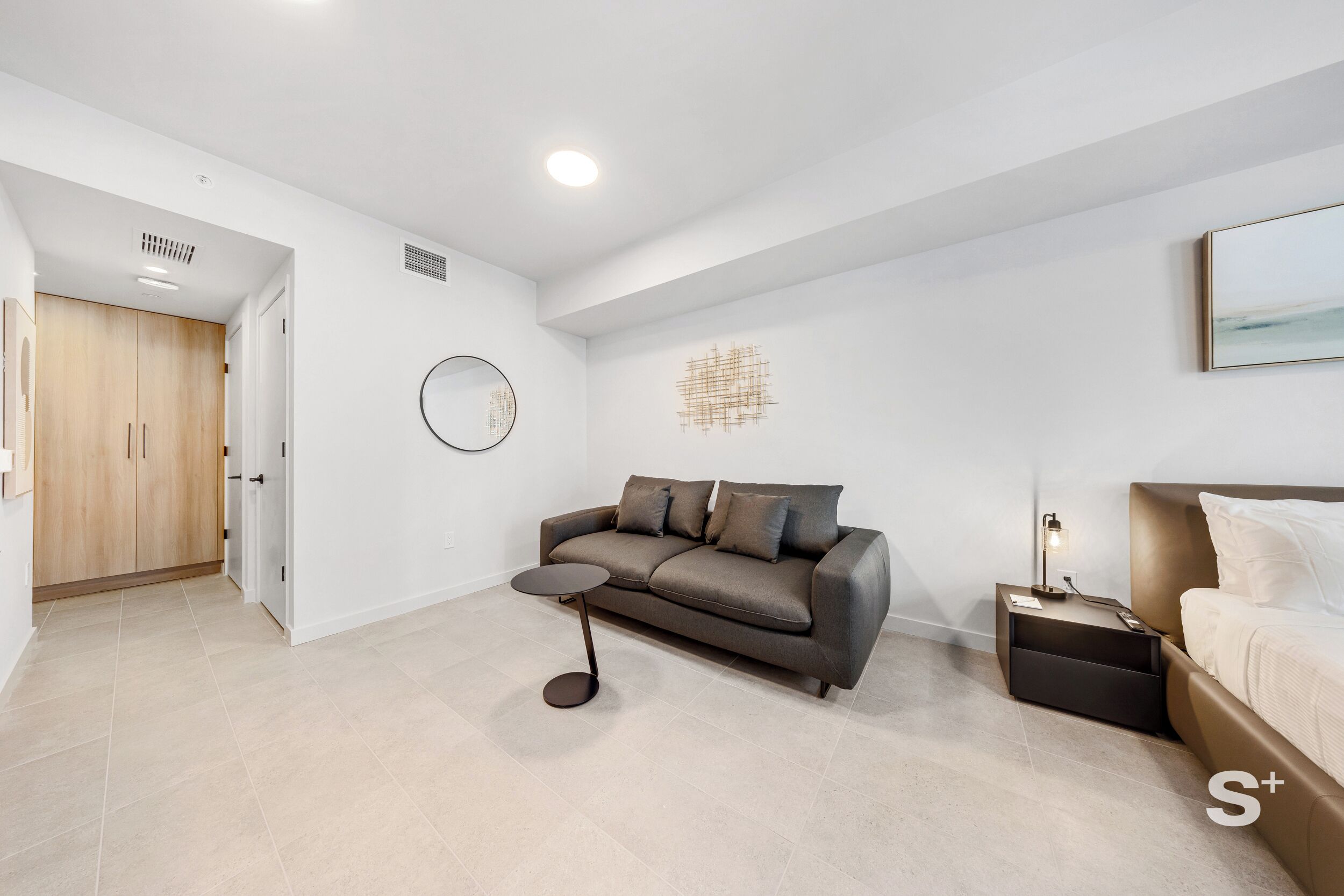 A modern living room with a sofa, a small round table, a bedside table with a lamp, and a bed.