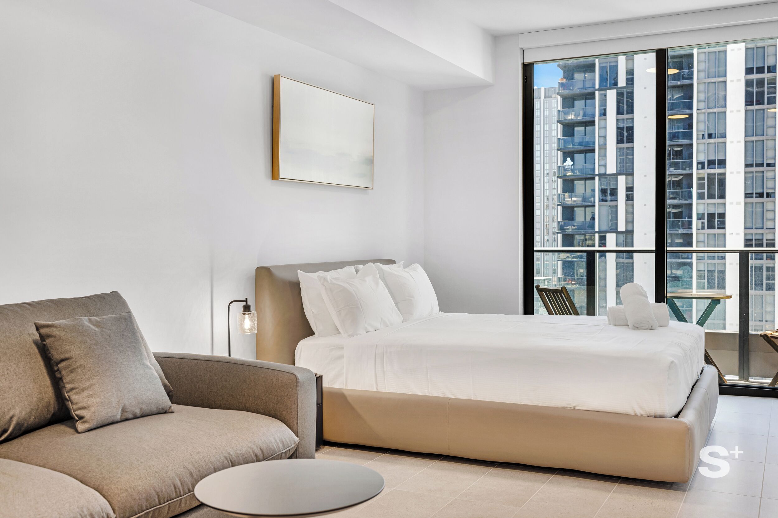 Une chambre d’hôtel moderne dotée d’un grand lit, d’un canapé, d’une table basse ronde et offrant une vue sur la ville grâce à de grandes fenêtres.