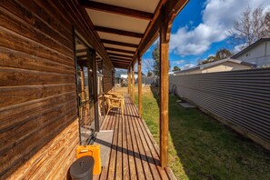 Una terraza de madera con un porche cubierto, mobiliario exterior y una valla metálica al fondo.