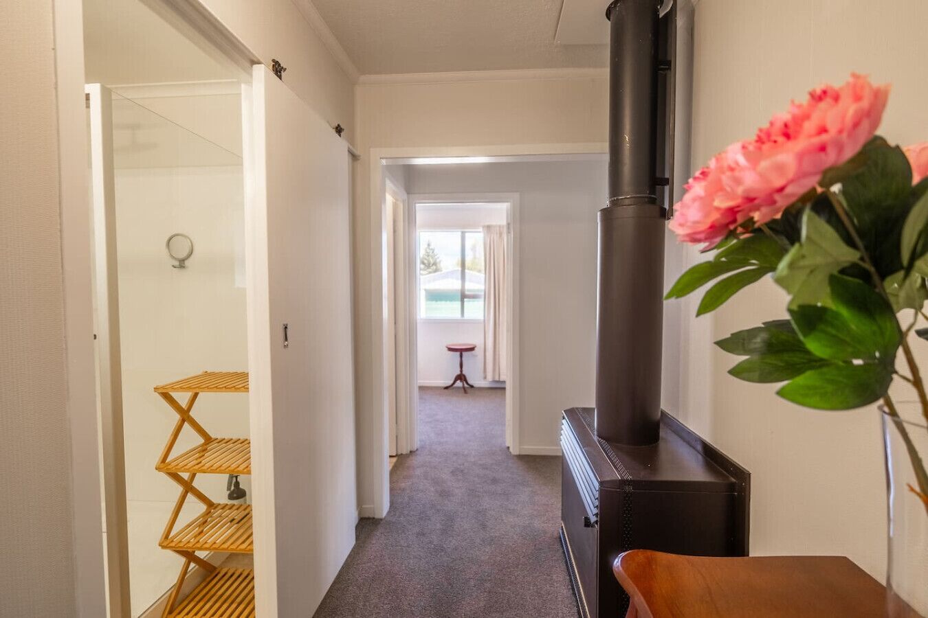 A hallway with a black wood-burning stove, a wooden shelf unit, and a pink flower in a vase.