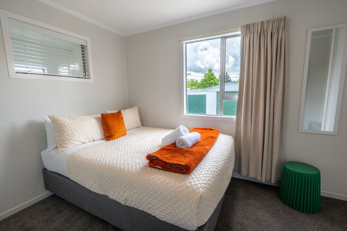 A hotel room with a bed, a window with curtains, a green stool, and a mirror on the wall.
