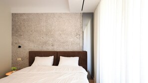 A modern bedroom with a large bed, a concrete accent wall, and a view of a cityscape through sheer curtains. - One Bedroom Apartment with City and Mountain View (Tiranë)