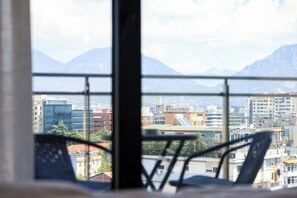 A view from a window with a metal railing, overlooking a cityscape with buildings and mountains in the background. - One Bedroom Apartment with City and Mountain View (Tiranë)