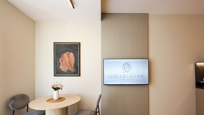 A reception area with a round table, two chairs, a framed artwork, and a television displaying 'Giselles Escape'. - One Bedroom Apartment with City and Mountain View (Tiranë)