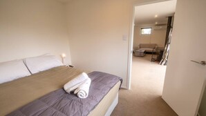 Ett hotellrum med en säng, en prydligt vikt handduk och en dörr som leder till ett vardagsrum med en soffa och en tv.