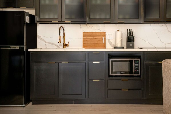 A modern kitchen with dark cabinetry, a stainless steel refrigerator, a built-in microwave, and a wooden cutting board on the countertop.