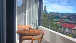 Ein Balkon mit Holztisch und Stühlen, einer kleinen Tischlampe und Blick auf Gebäude und Bäume.