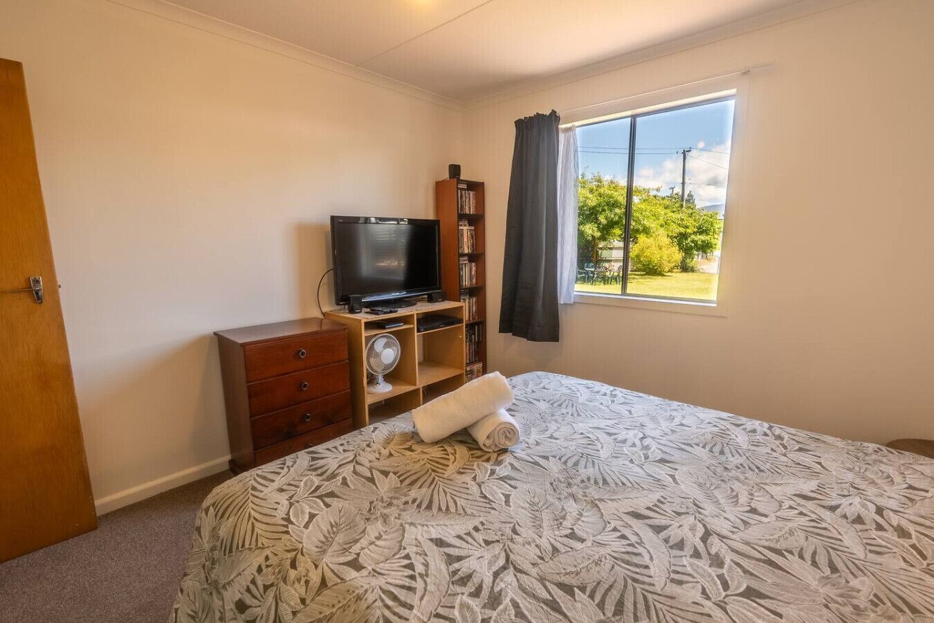 A bedroom with a bed, a television, a bookshelf, a fan, and a window with a view of greenery.