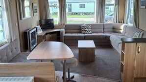 A cozy RV interior with a sofa, a table, a TV, and a view of a green mobile home outside. - Glasyln 37 (Porthmadog)