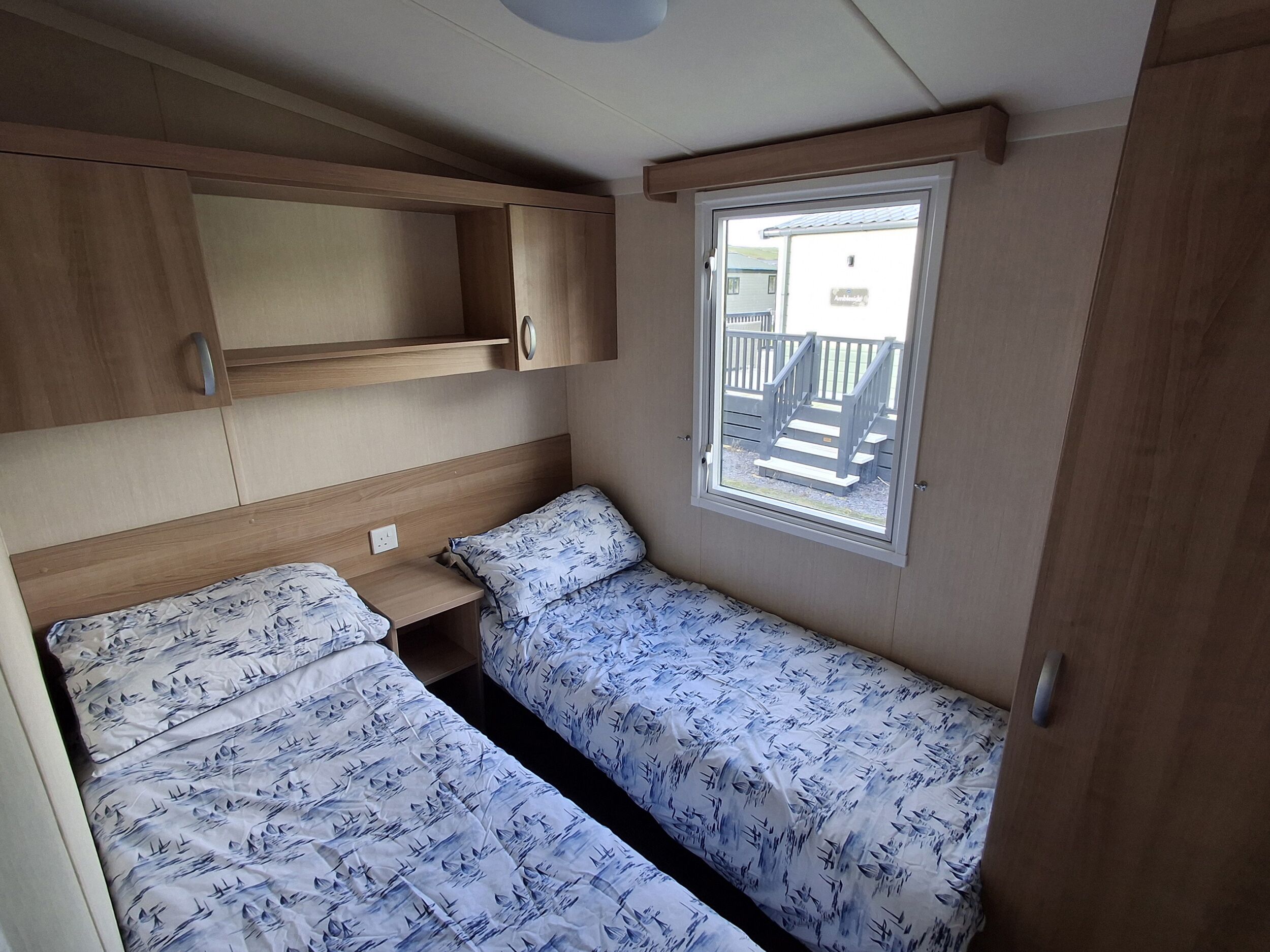 A compact RV bedroom with two beds, a wooden headboard, a window with a view of a deck, and built-in storage cabinets.