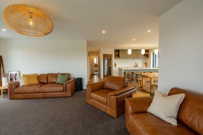 A spacious living room with a brown leather sofa set, a coffee table, and a large woven light fixture. A kitchen with wooden cabinets and a dining area is visible in the background. - Modern Glenfield - Private & Spacious (Twizel)