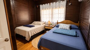 Habitación con dos camas, una mesita de noche con lámpara, ventana con cortinas y suelo de madera.