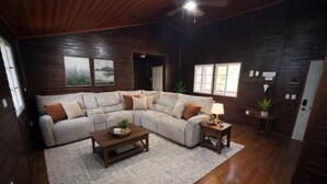 Una sala de estar acogedora con un sofá seccional, una mesa de centro y una mesita de noche con una lámpara. Hay dos ventanas que proporcionan luz natural y un ventilador de techo.