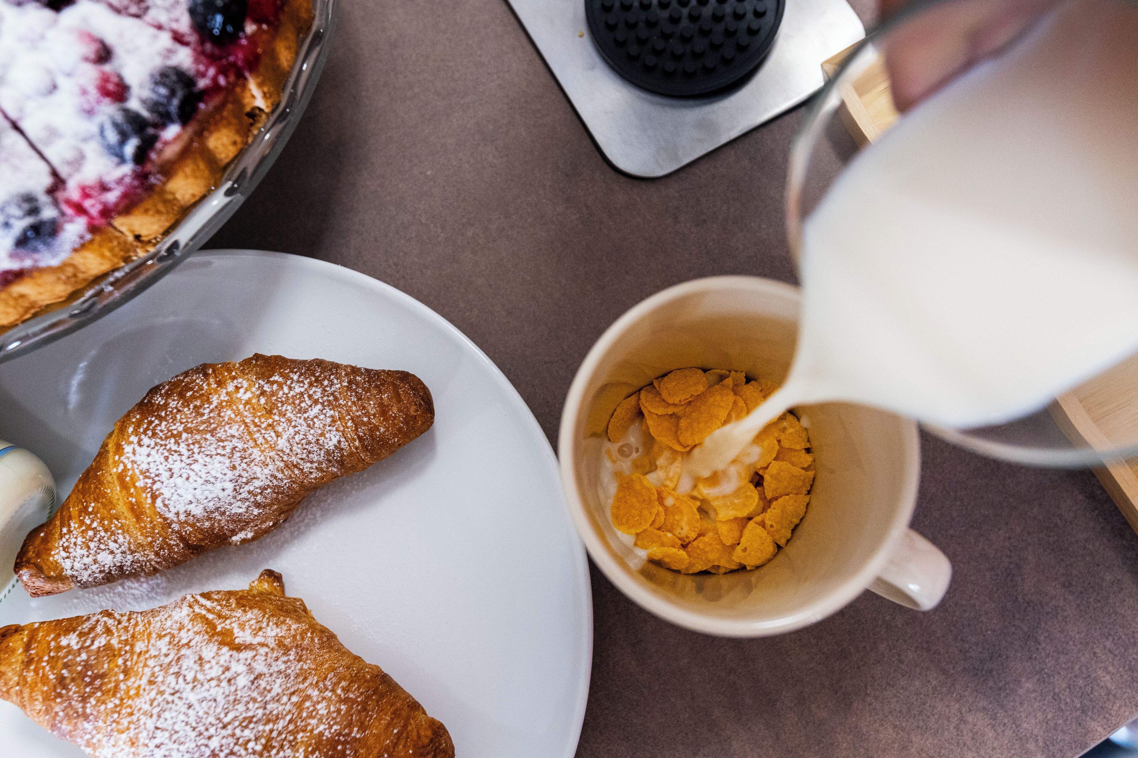 Un desayuno con pastelería, un tazón de cereal y una taza siendo servida con leche.
