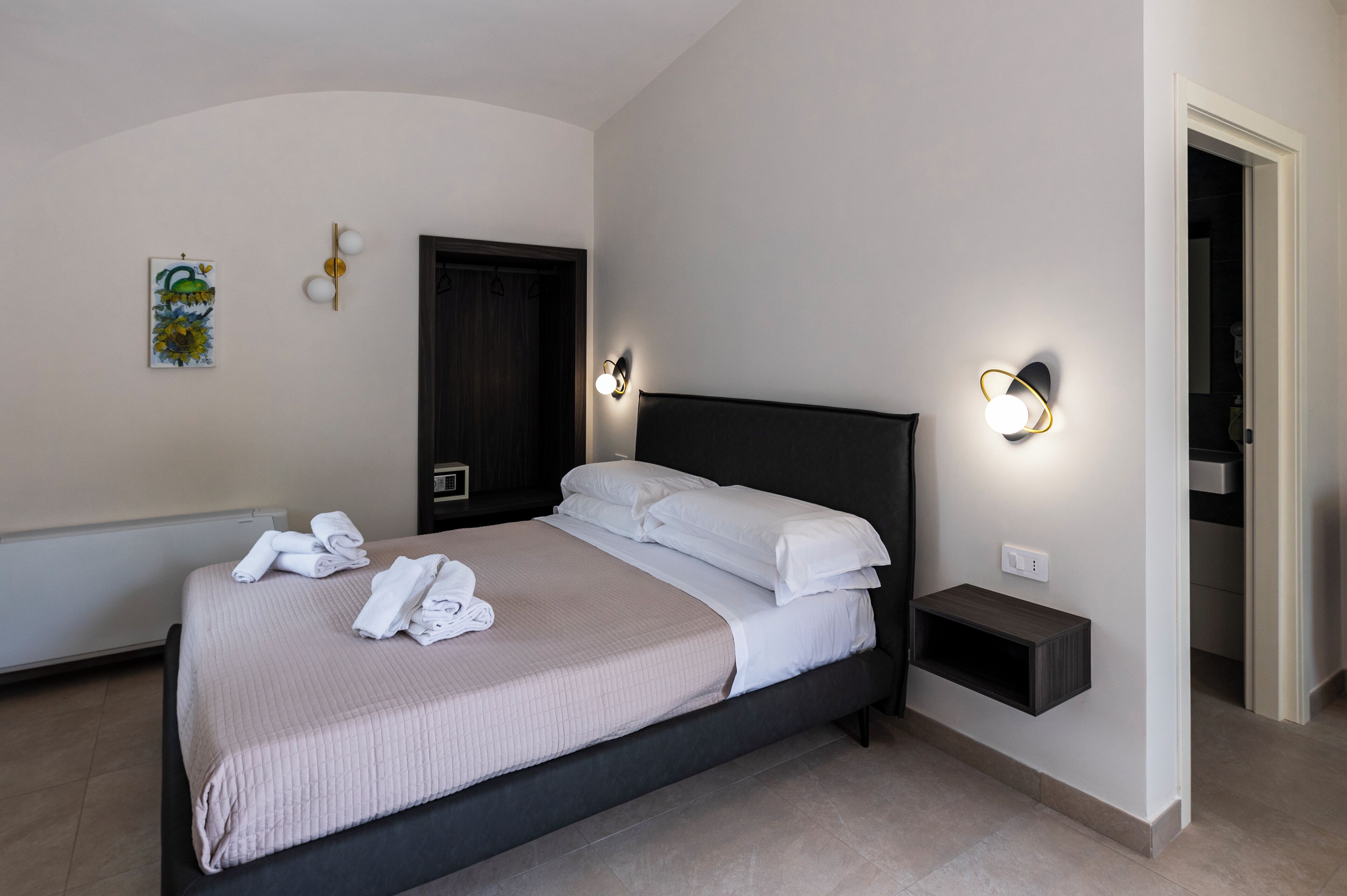 Una cama bien hecha con sábanas blancas, una mesita de noche con lámpara y un estante en la pared.