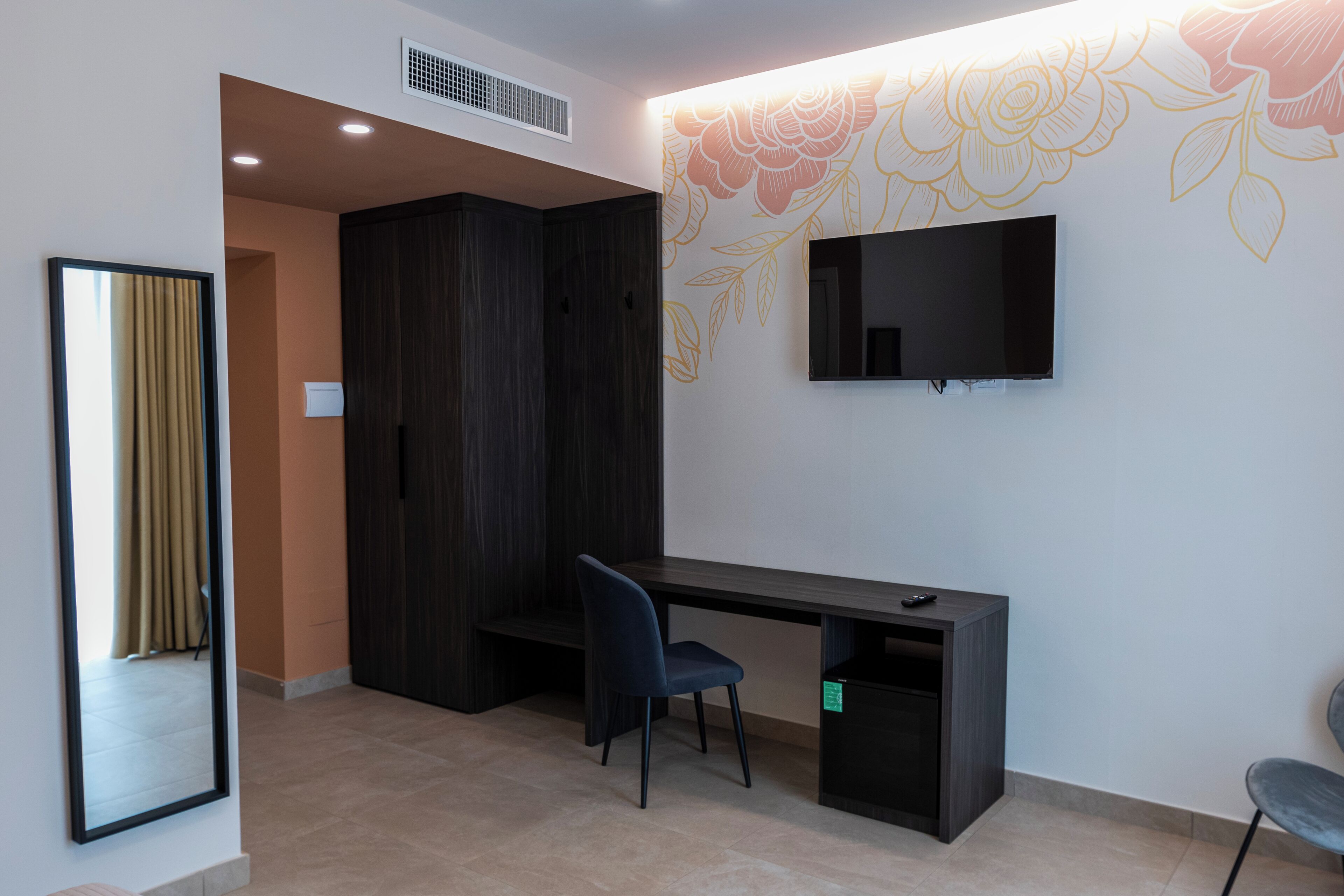 A room with a dark wood wardrobe, a desk with a chair, a flat-screen TV mounted on the wall, and a mirror on the left.