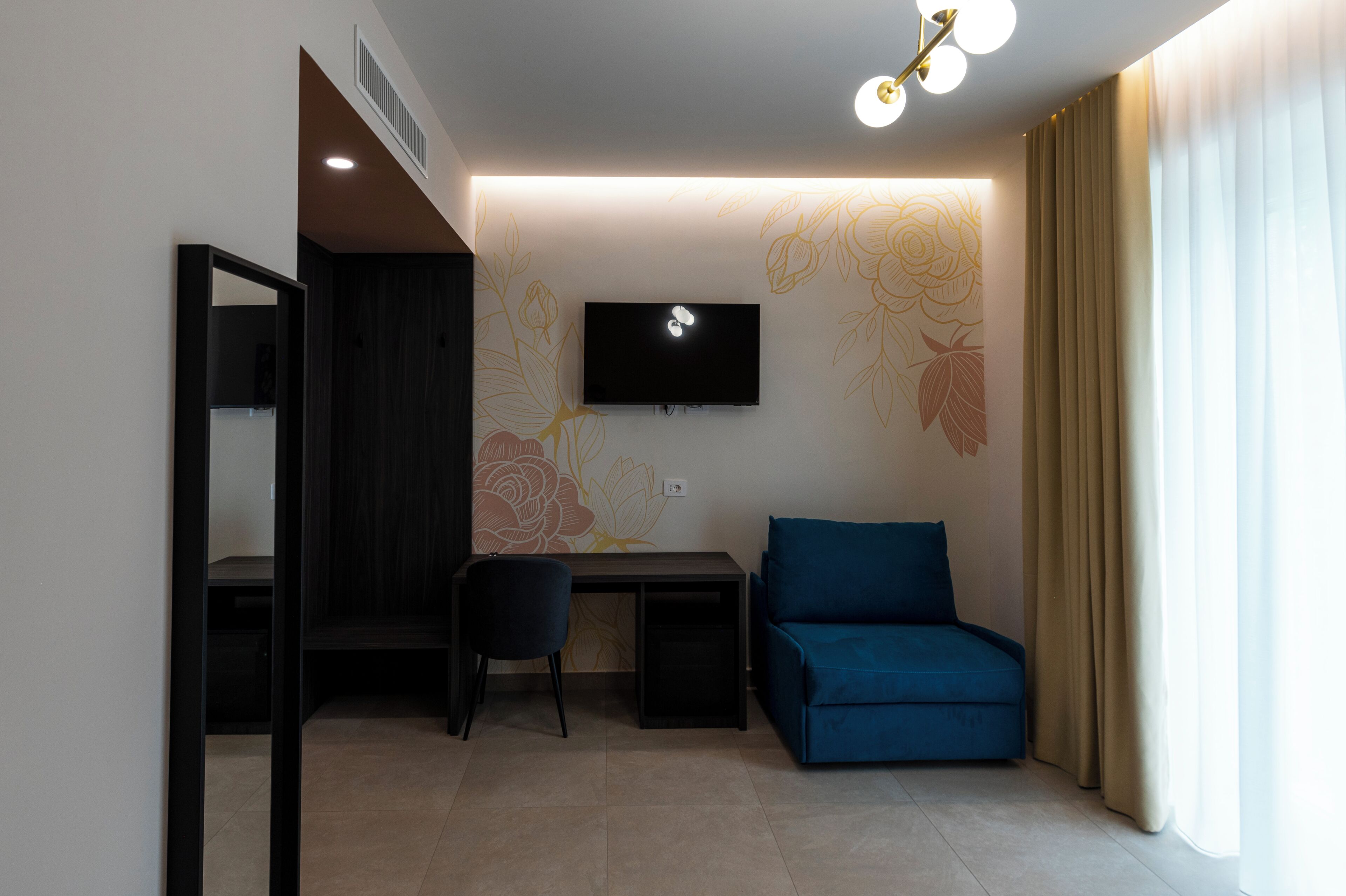 Ein modernes Zimmer mit einem flachbildfernseher an der Wand, einem Schreibtisch mit Stuhl, einem blauen Sofa und einem großen Fenster mit Vorhängen.