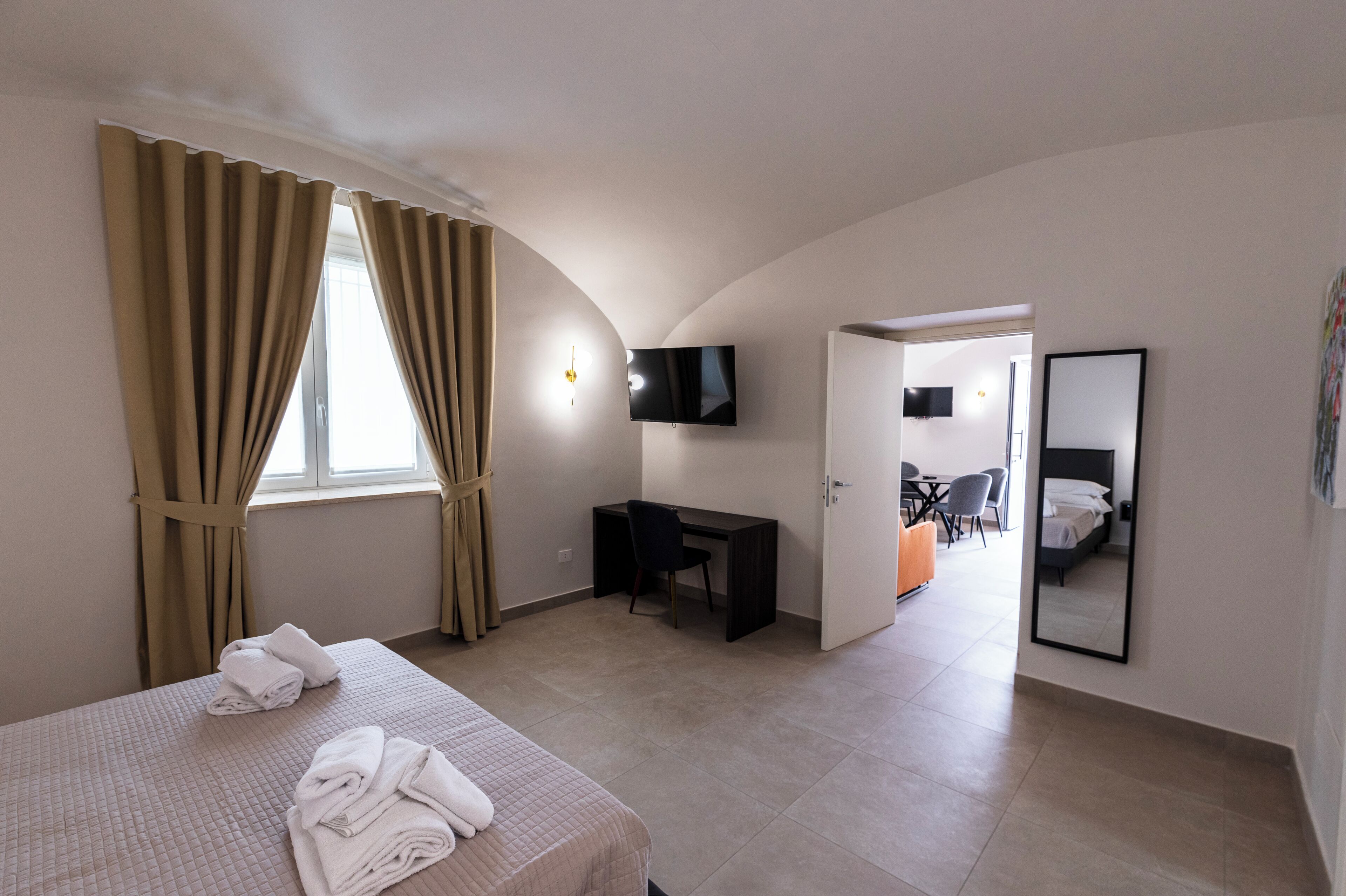 Habitación de hotel con cama, escritorio, silla, televisor, espejo y puerta que da a otra habitación.