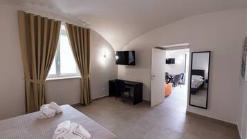 Habitación de hotel con cama, escritorio, silla, televisor, espejo y puerta que da a otra habitación.