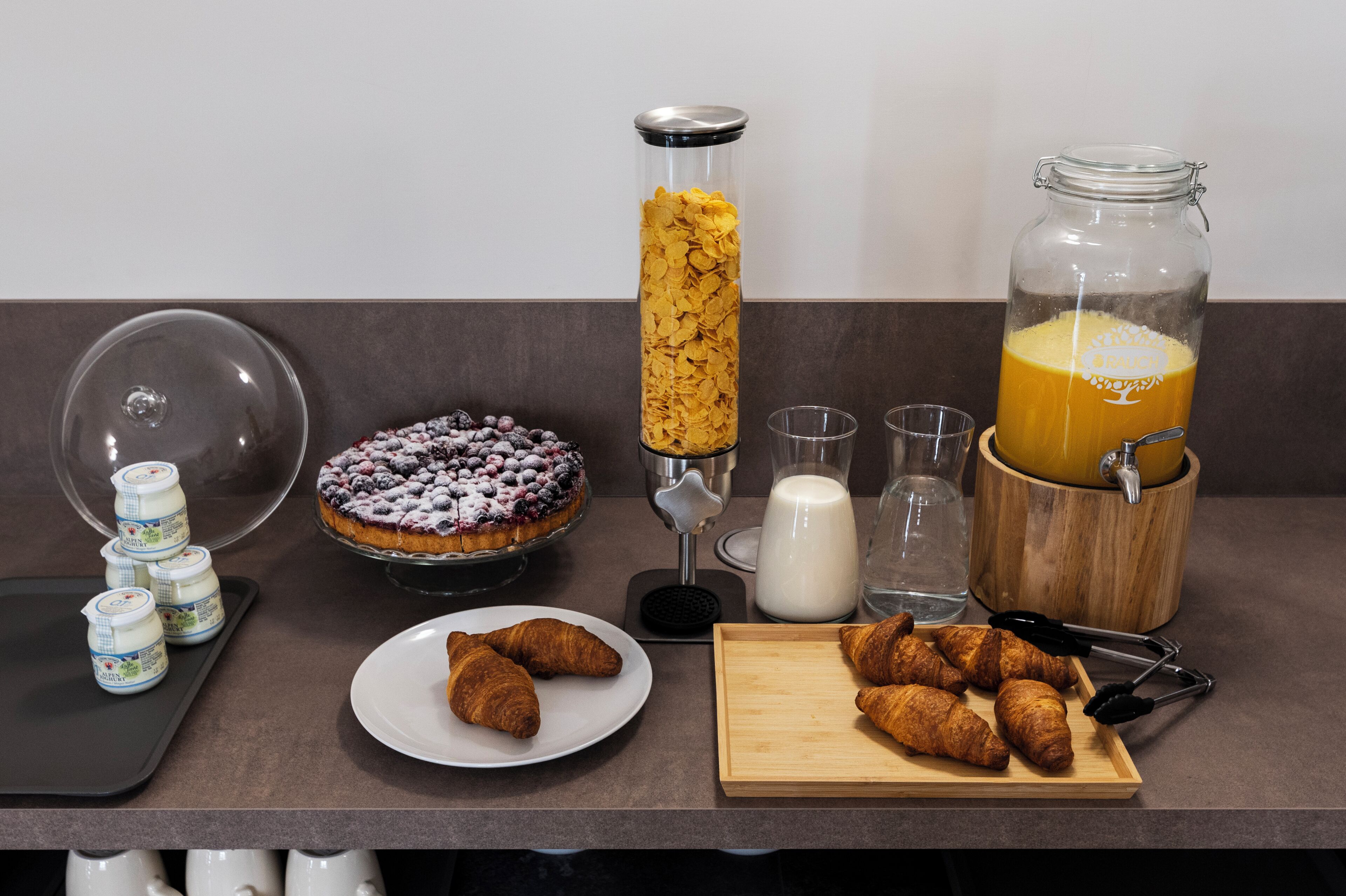 A breakfast setup with a fruit tart, croissants, a juice dispenser, milk, and yogurt containers.