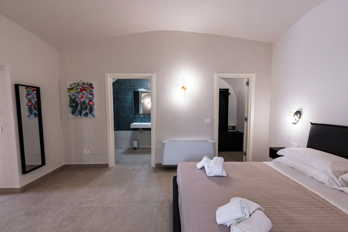 A hotel room with a bed, a bathroom with a mirror, a radiator, and a painting on the wall.