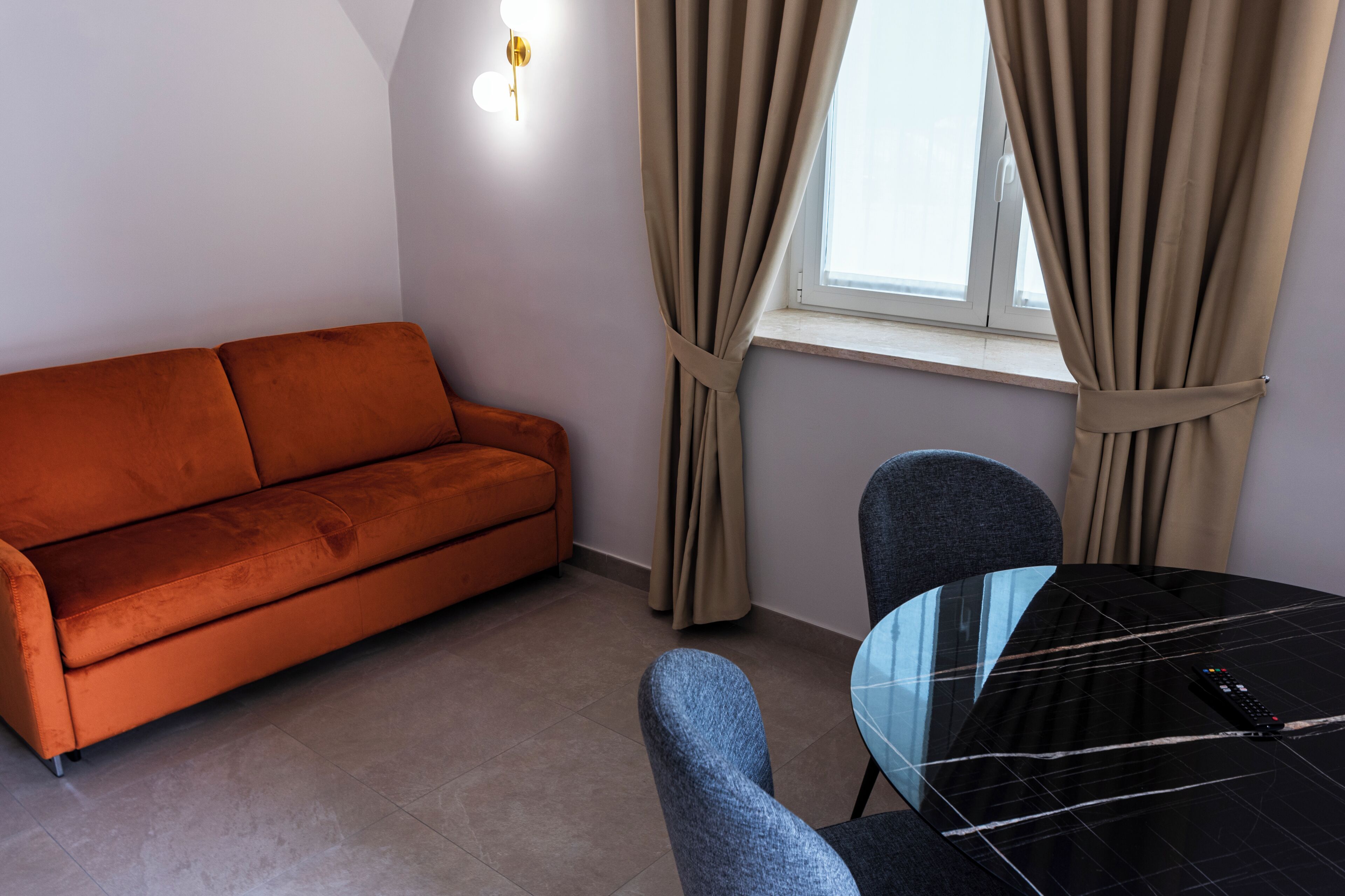 Ein Zimmer mit einer Couch, einem Tisch mit Stühlen, einem Fenster mit Vorhängen und einer Wandleuchte.
