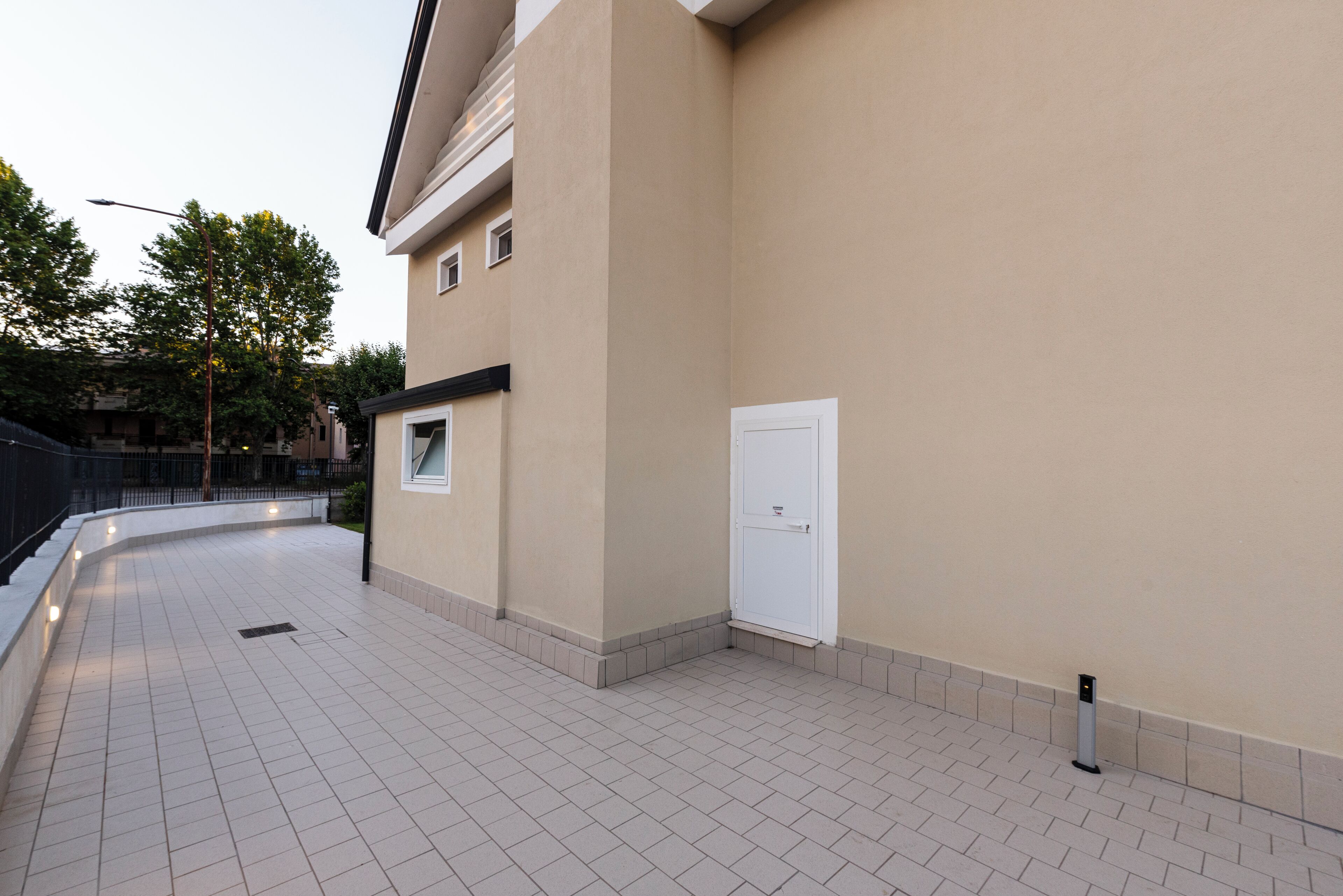 Une façade d’immeuble avec un trottoir dallé, une porte blanche et une petite fenêtre.