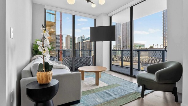 Ein modernes Wohnzimmer mit Sofa, Sessel und kleinem Tisch, großen Fenstern mit Blick auf die Stadt und einem an der Wand montierten Flachbildfernseher.