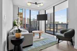 Un salon moderne avec un canapé, un fauteuil et une petite table, de grandes fenêtres offrant une vue sur la ville, et un téléviseur à écran plat fixé au mur.