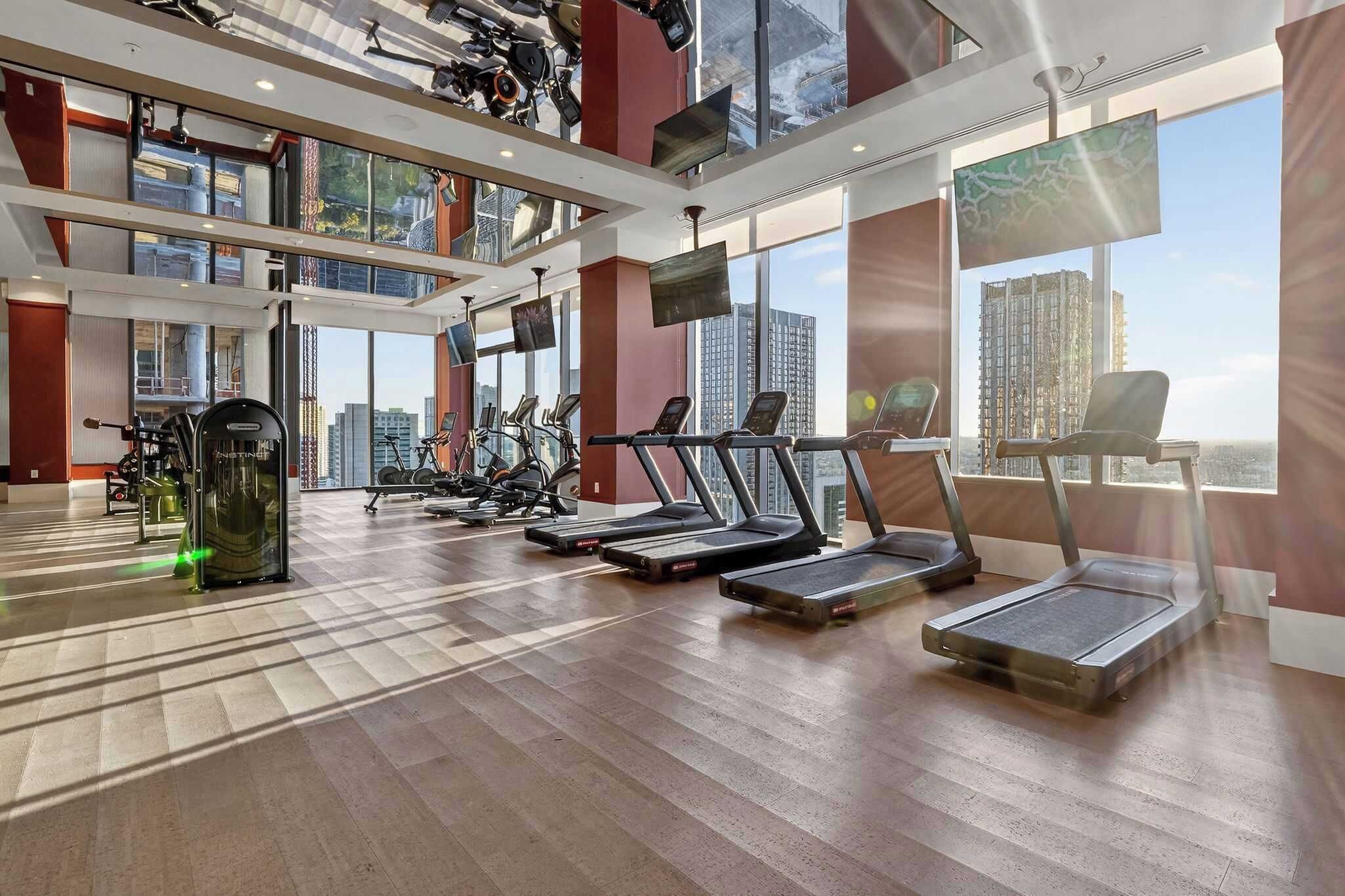 A modern gym with treadmills, exercise bikes, and weight machines, large windows with a city view, and a high ceiling with recessed lighting.
