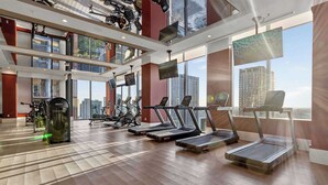 Ein modernes Fitnessstudio mit Laufbändern, Ergometer und Krafttrainingsgeräten, großen Fenstern mit Blick auf die Stadt und einer hohen Decke mit eingelassener Beleuchtung.