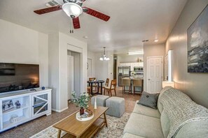 A living room with a TV, a ceiling fan, a dining area with a table and chairs, and a kitchen in the background. - Modern 2BR Condo w/ Pool & Parking in Scottsdale (Scottsdale)