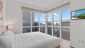 Une chambre moderne avec un grand lit, une vue sur la ville grâce à de grandes fenêtres du sol au plafond et un téléviseur fixé au mur.