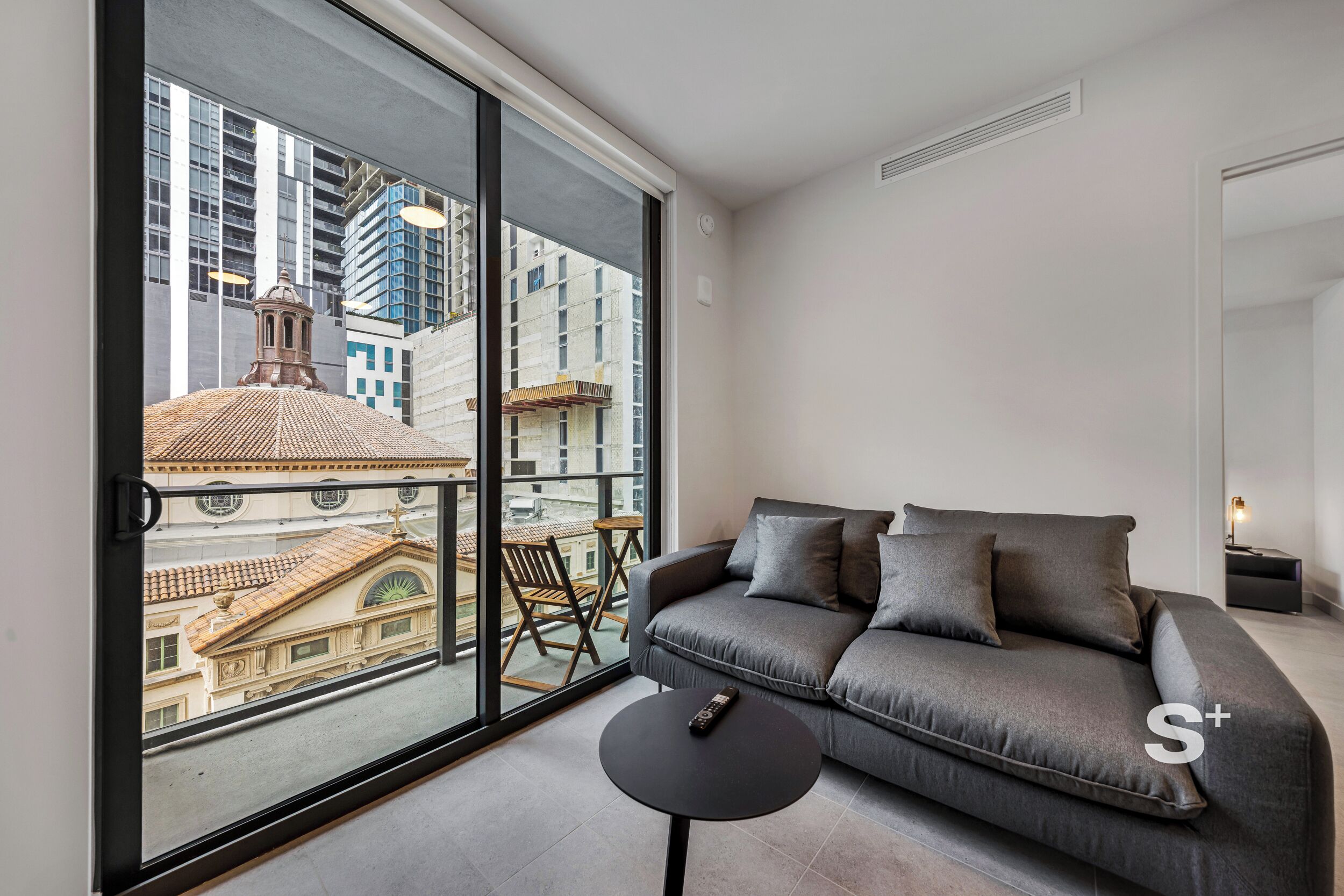 Ein modernes Wohnzimmer mit einem grauen Sofa, einem kleinen runden Tisch und einem großen Fenster mit Blick auf Stadtgebäude.