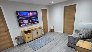 A modern living room with a flat-screen TV mounted on the wall, a grey rug, a wooden cabinet, a grey patterned sofa, and a wooden side table. - 2 Bed House Sleeps 5 -private Garden (Stirling)