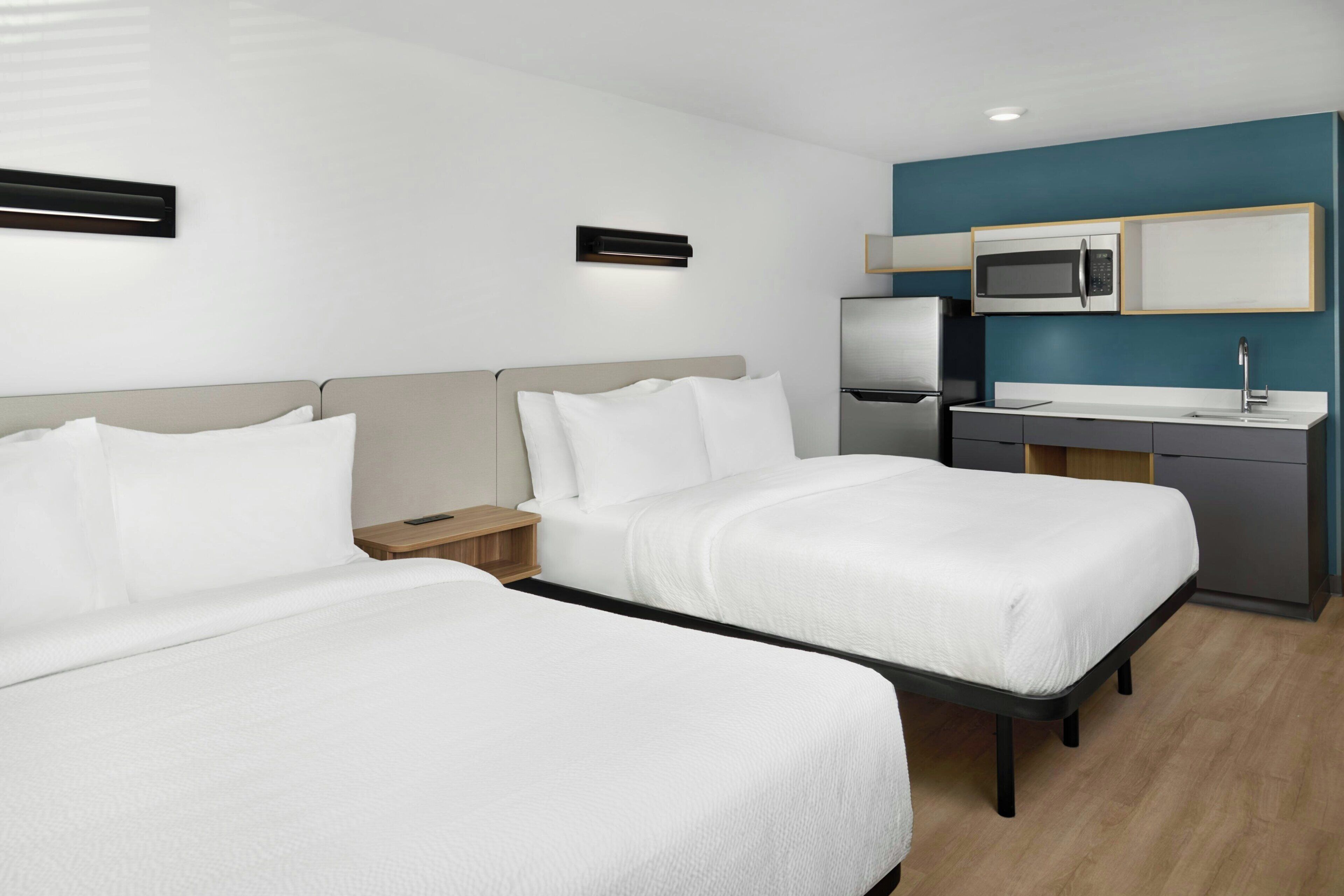 Een hotelkamer met twee bedden, een kitchenette en een klein nachtkastje.