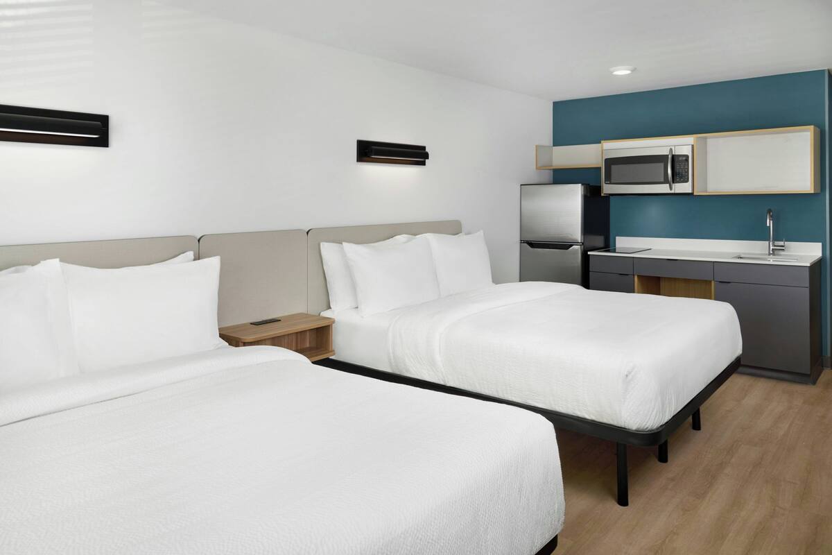 Een hotelkamer met twee bedden, een kitchenette en een klein nachtkastje.