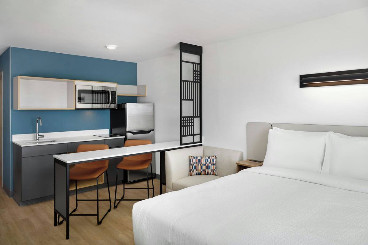 Een compacte hotelkamer met een kitchenette, een bed, een bureau en een stoel.