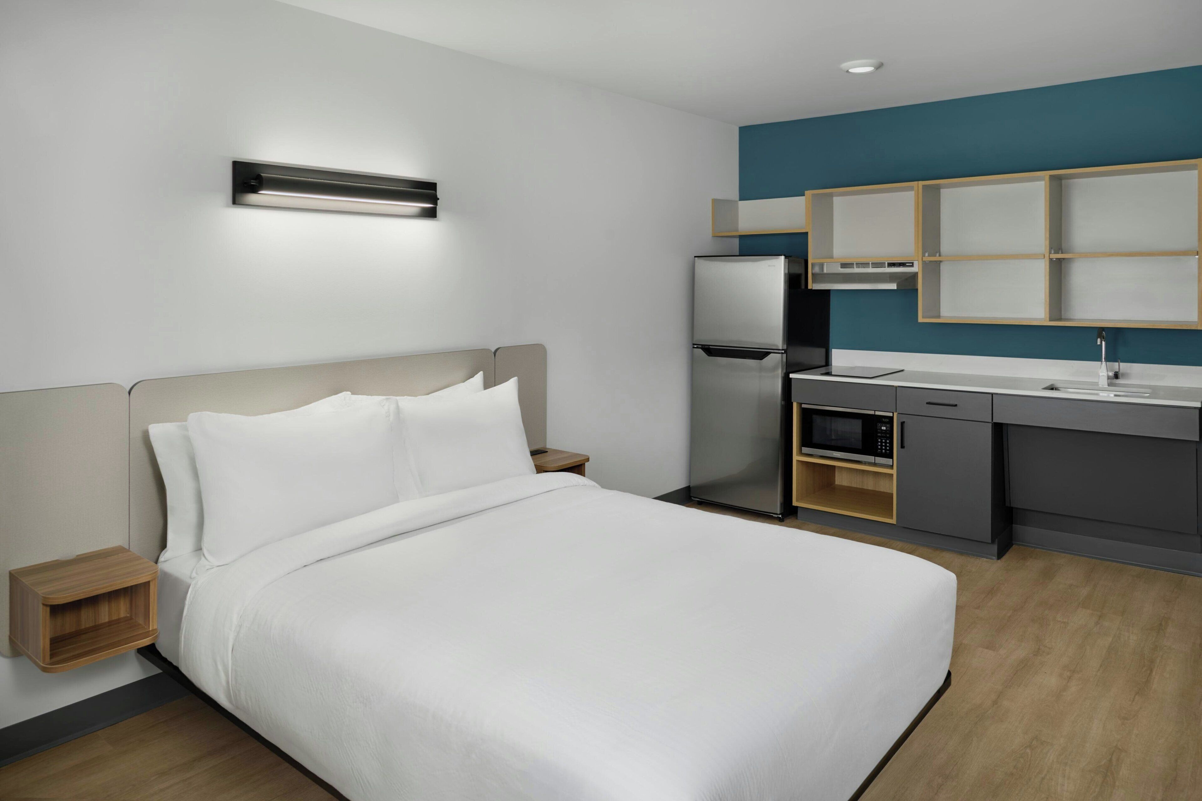 A modern hotel room with a bed, bedside table, built-in microwave, and a kitchenette with a refrigerator and sink.