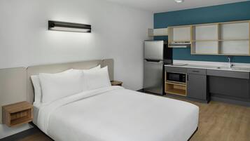 A modern hotel room with a bed, bedside table, built-in microwave, and a kitchenette with a refrigerator and sink.