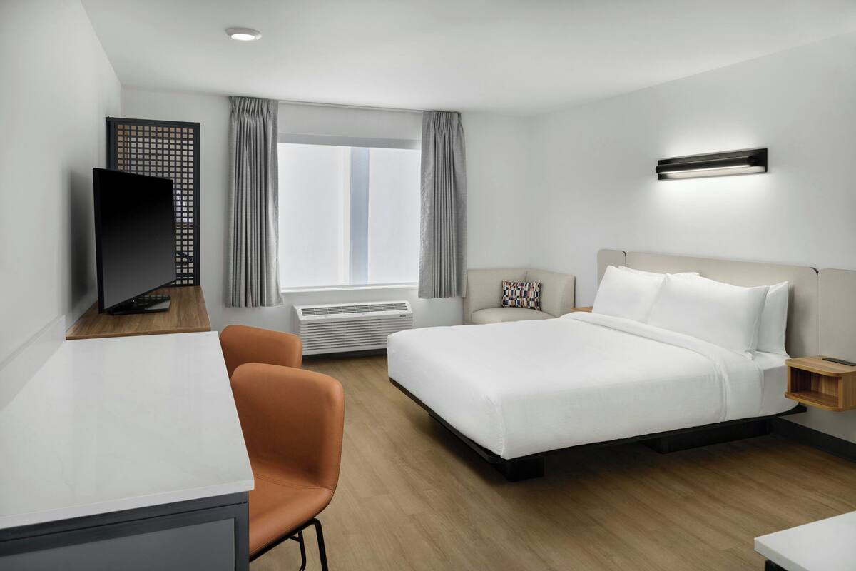 Een hotelkamer met een bed, een bureau, een televisie, een raam met gordijnen en een wandlamp.