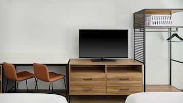 A hotel room with a TV, wooden desk, two chairs, a bed, and a laundry basket.