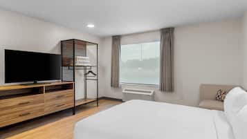 A hotel room with a bed, a TV, a wooden dresser, a hanging clothes rack, and a window with curtains.