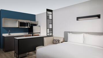 Una habitación de hotel moderna con una cama, un escritorio con microondas, una kitchenette pequeña y un panel decorativo en la pared.