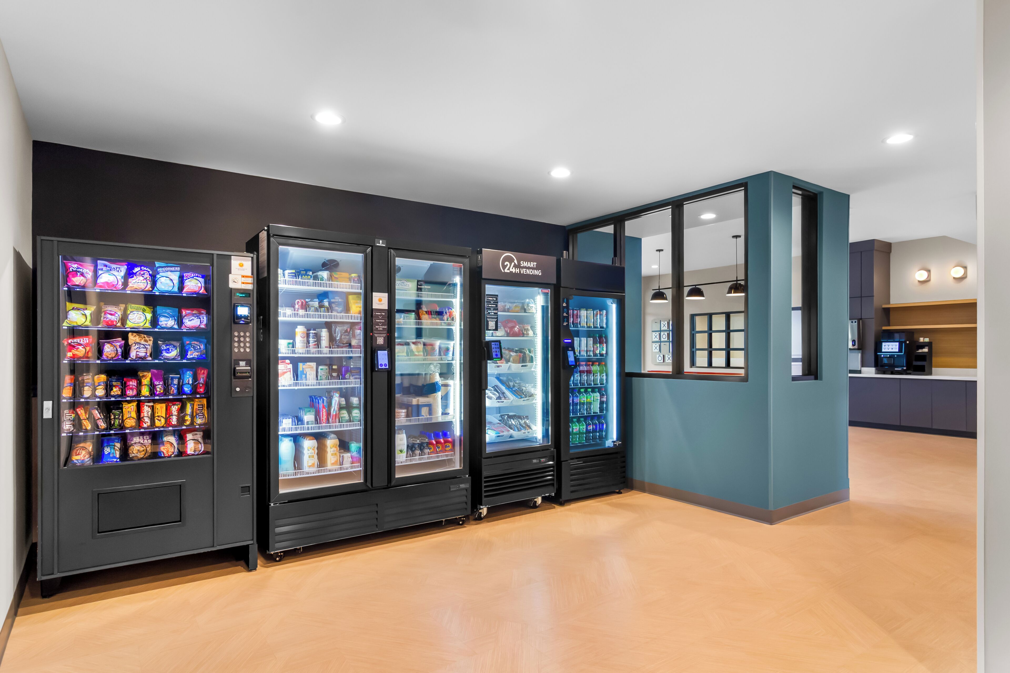 Una hilera de máquinas expendedoras con aperitivos y bebidas, una pared color verde azulado y un piso de madera.