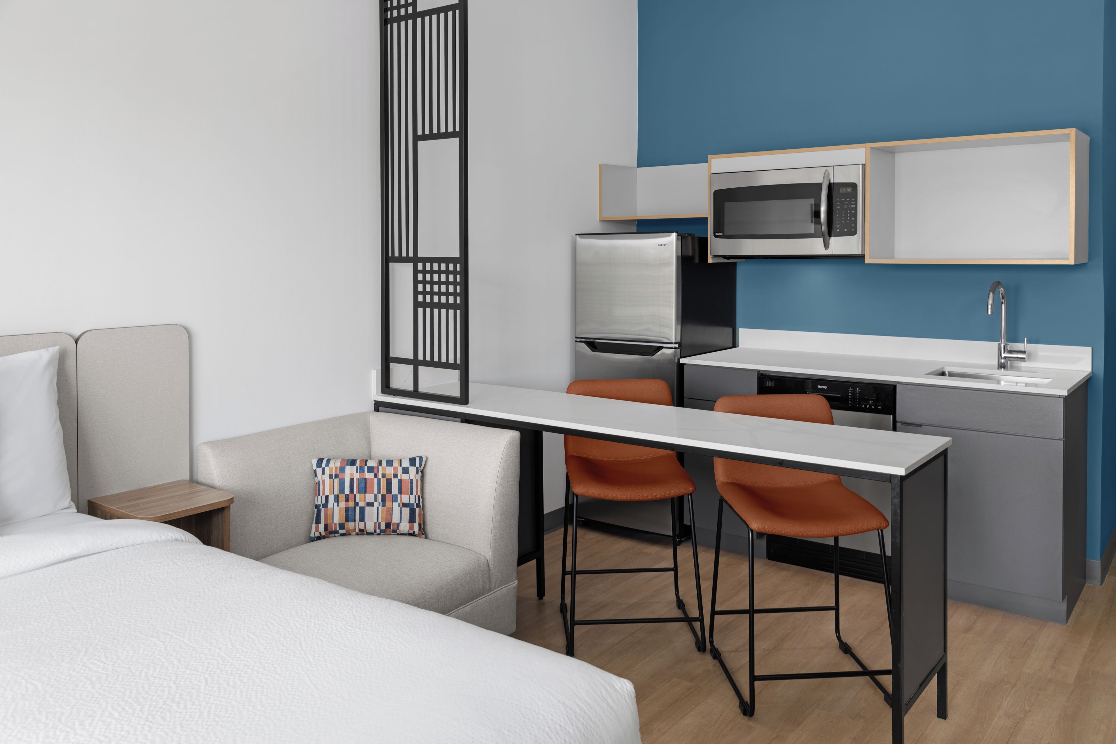 Une chambre d’hôtel compacte comprenant un lit, un petit bureau, deux chaises, une kitchenette et un four à micro-ondes.