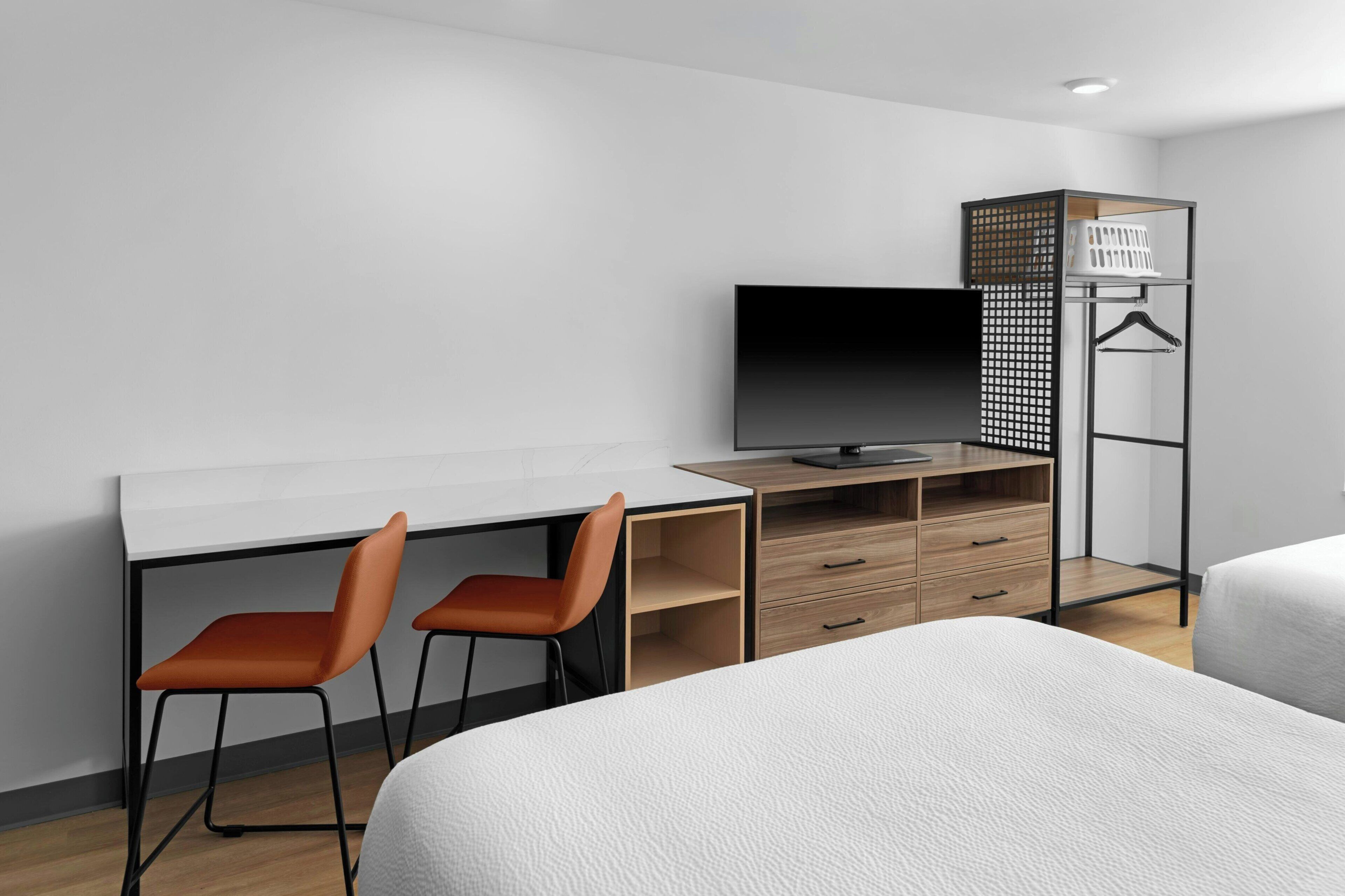 A hotel room with a bed, a desk with two chairs, a TV, a wardrobe, and a hanging rack.