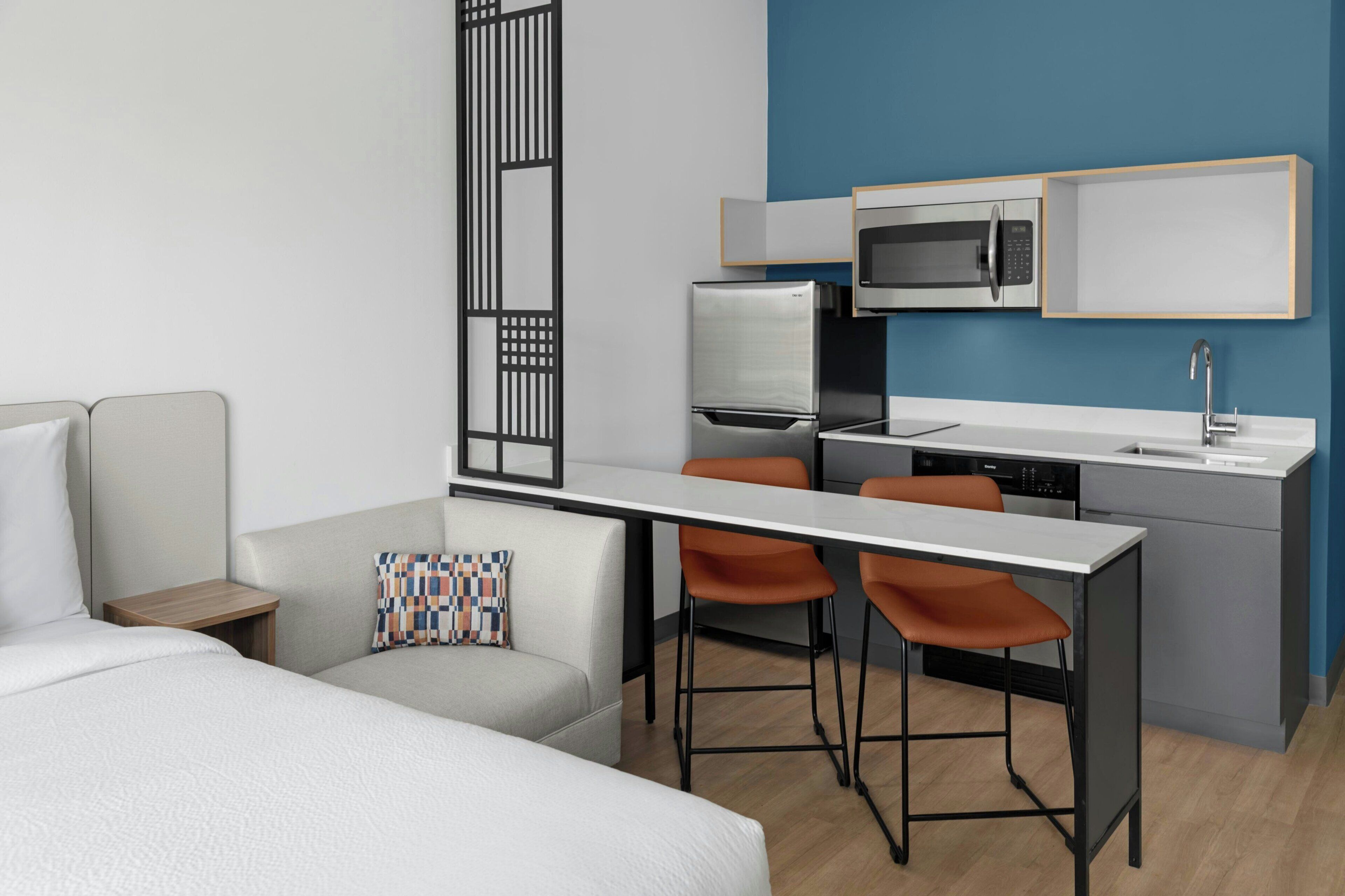 A modern hotel room with a bed, a desk, two chairs, a microwave, a refrigerator, and a dishwasher.