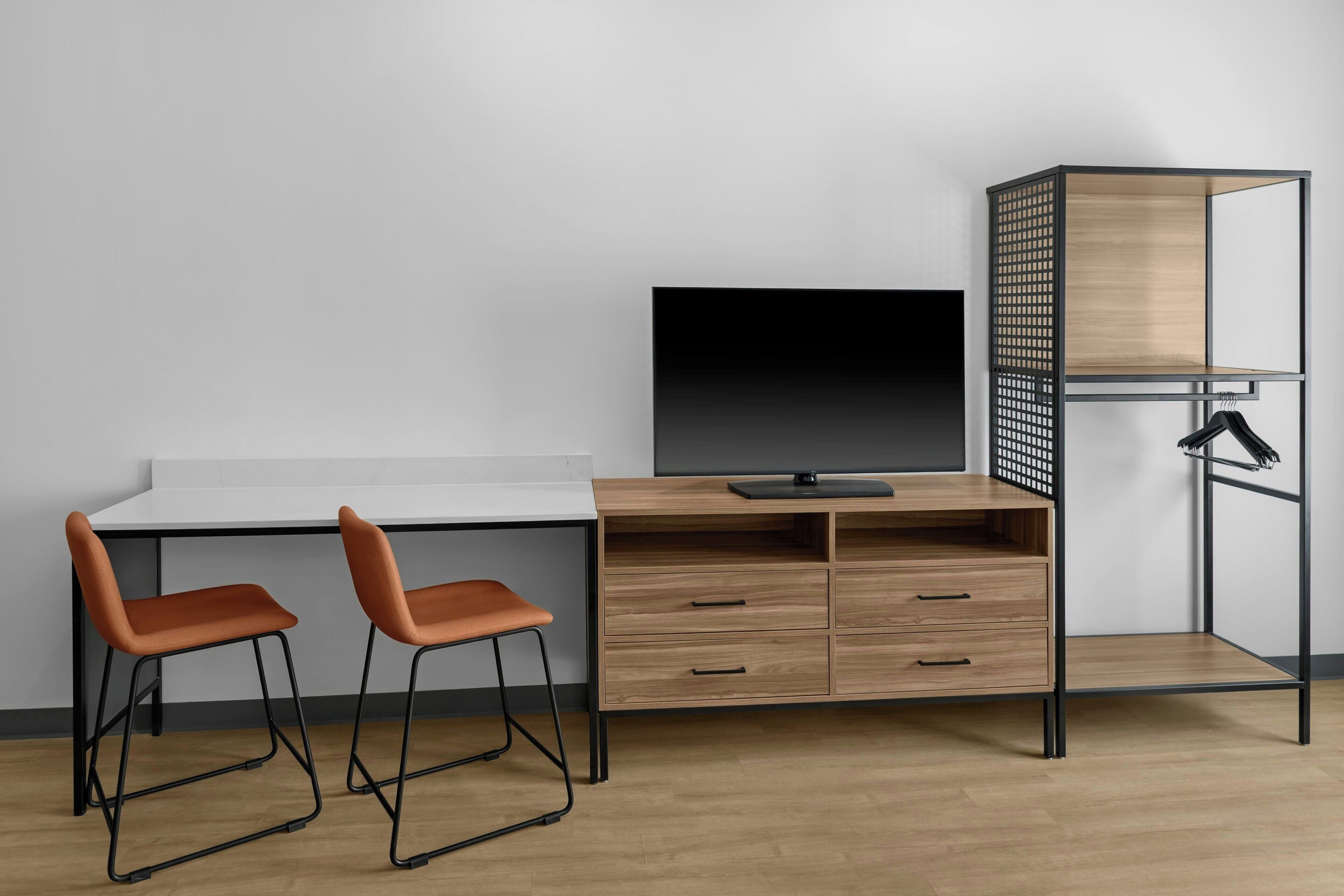 Un espacio de trabajo moderno con un escritorio blanco, dos sillas marrones, un monitor negro y una unidad de estanterías de madera con estructura metálica negra.